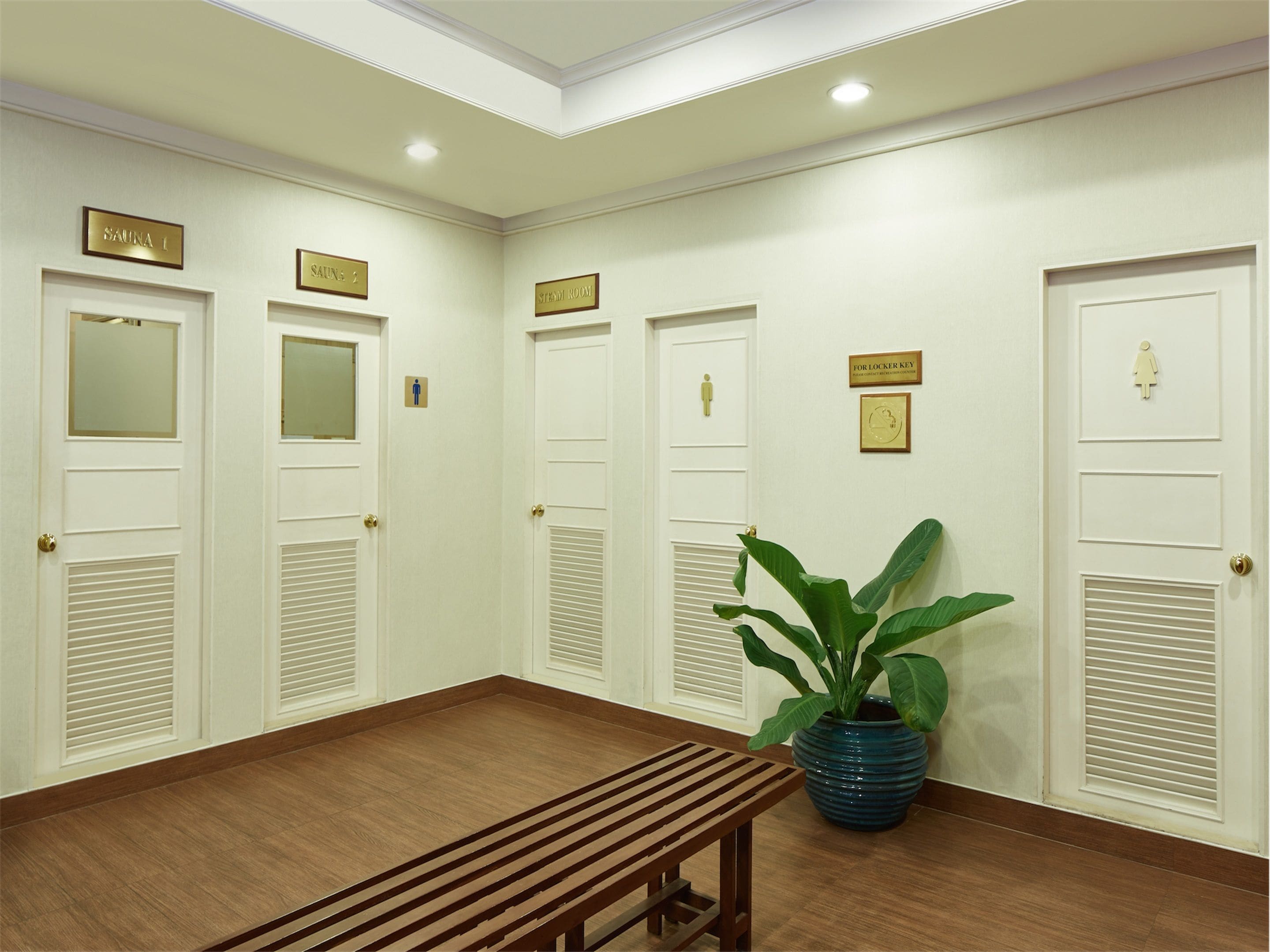 Une chambre d’hôtel avec un banc en bois, une plante en pot et des portes blanches sur lesquelles sont indiqués le numéro de la chambre et les équipements disponibles.