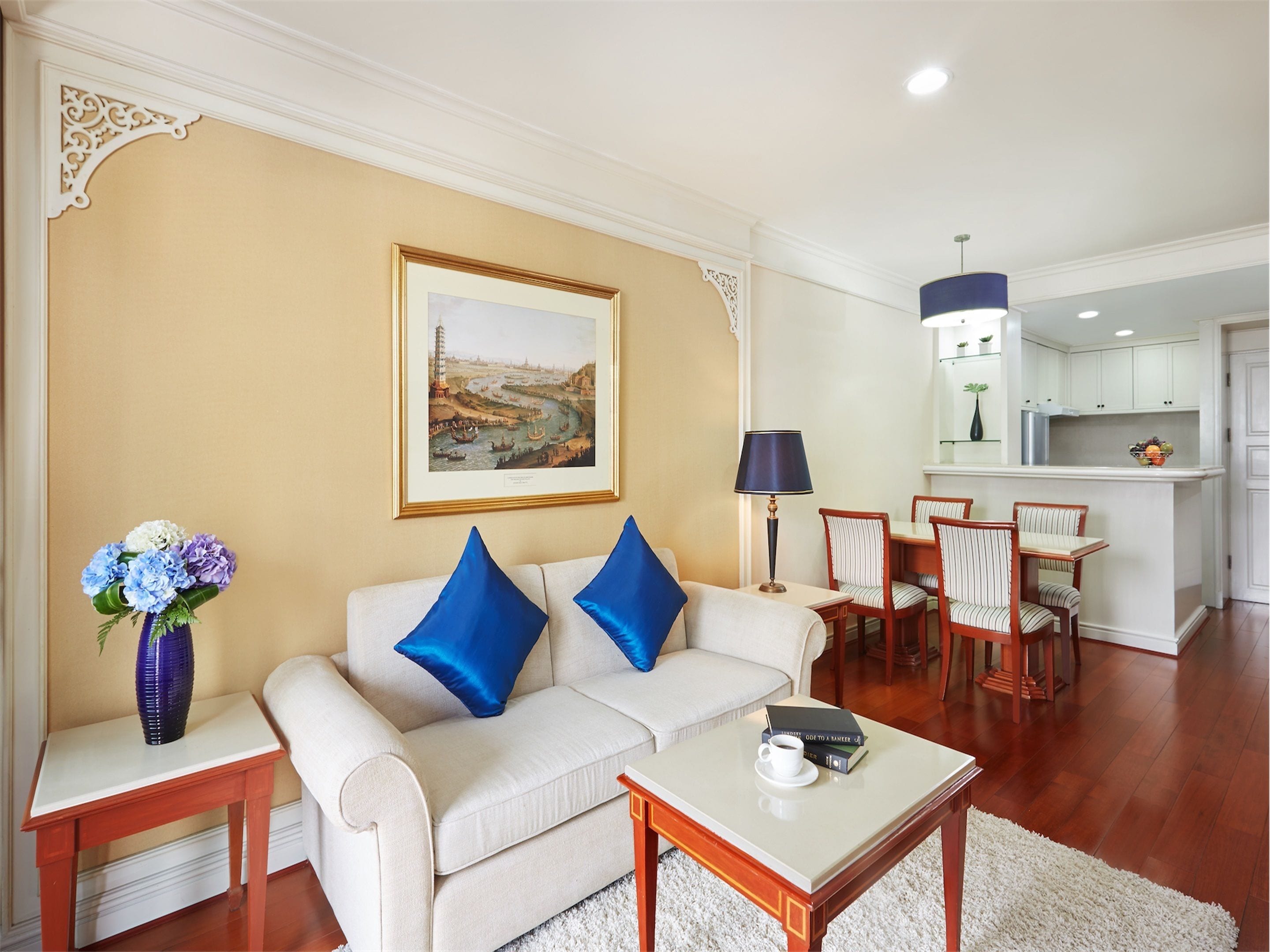 A living room with a beige sofa, two blue pillows, a coffee table with a cup and books, a framed landscape painting, a vase with flowers, and a dining area with striped chairs and a table.