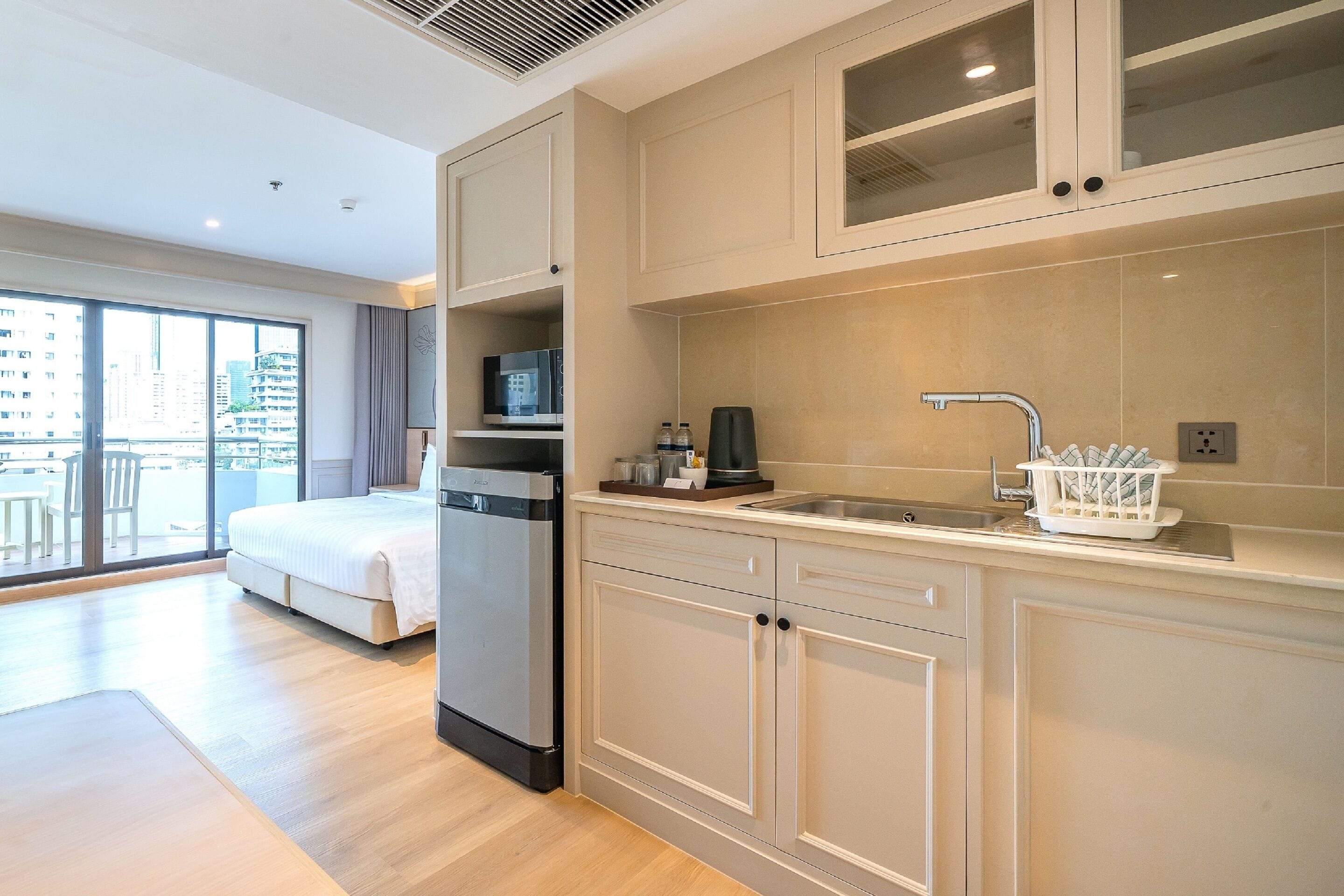 A modern hotel room with a kitchenette, a bed, a large window with a city view, and a balcony.