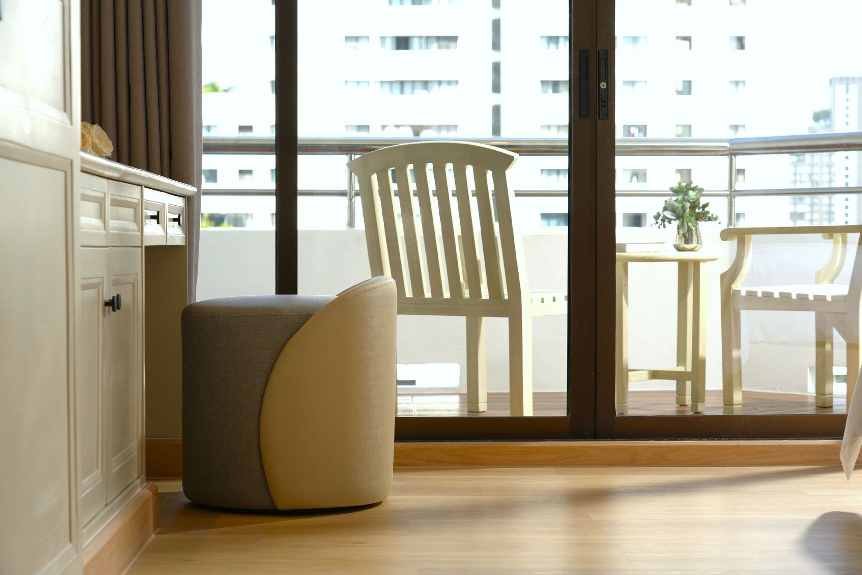 A modern room with a wooden floor, a white chair, a small table with a plant, and a view of a building through large windows.