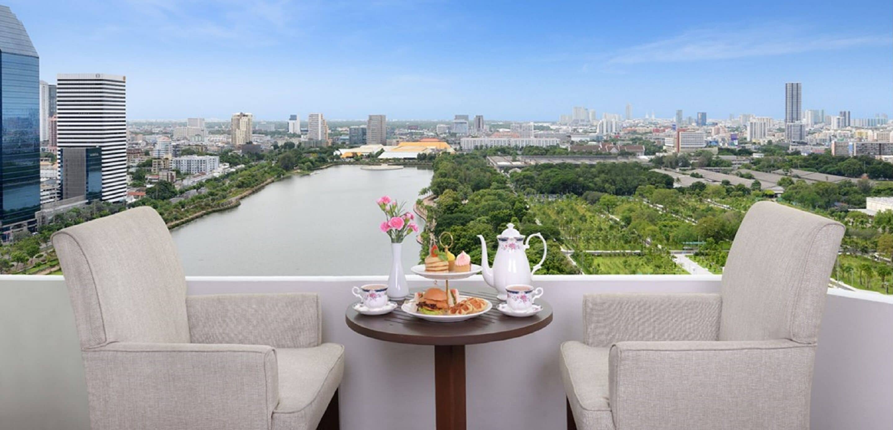 A balcony with two chairs, a table with a teapot, cups, and food, overlooking a cityscape and a river.
