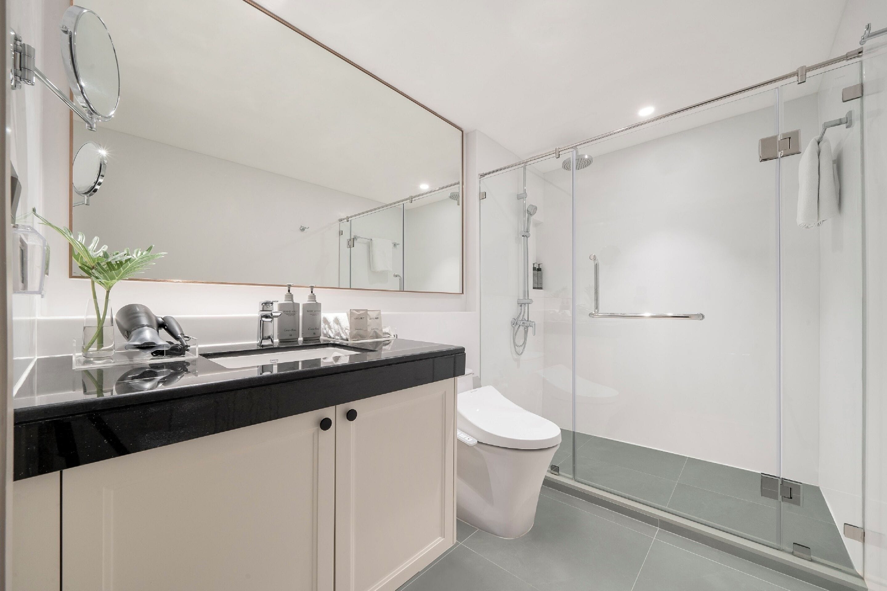 Un baño moderno con encimera negra, armarios blancos, inodoro blanco y zona de ducha con mampara de vidrio.