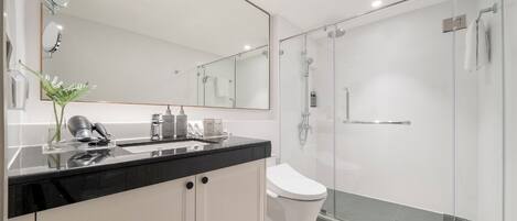 Un baño moderno con encimera negra, armarios blancos, inodoro blanco y zona de ducha con mampara de vidrio.