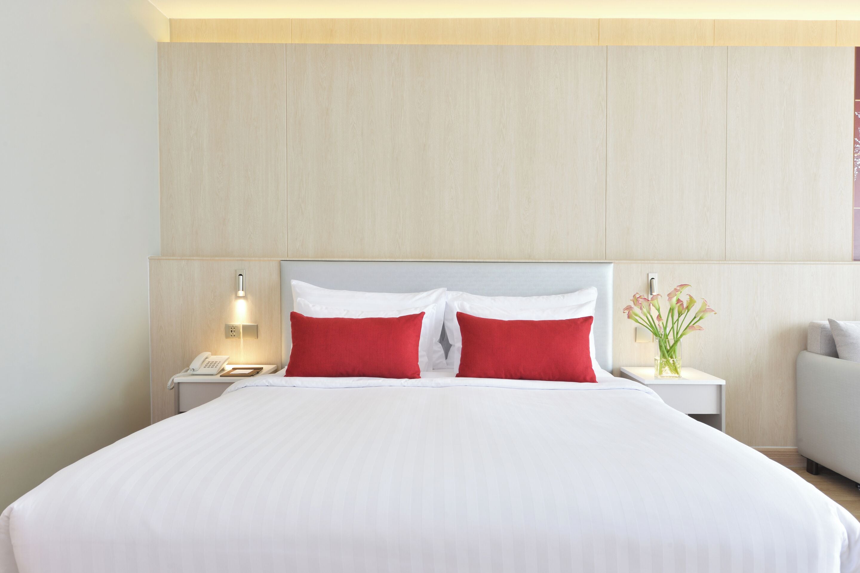 A hotel room with a large bed, two red pillows, a bedside table with a lamp, and a vase with flowers.