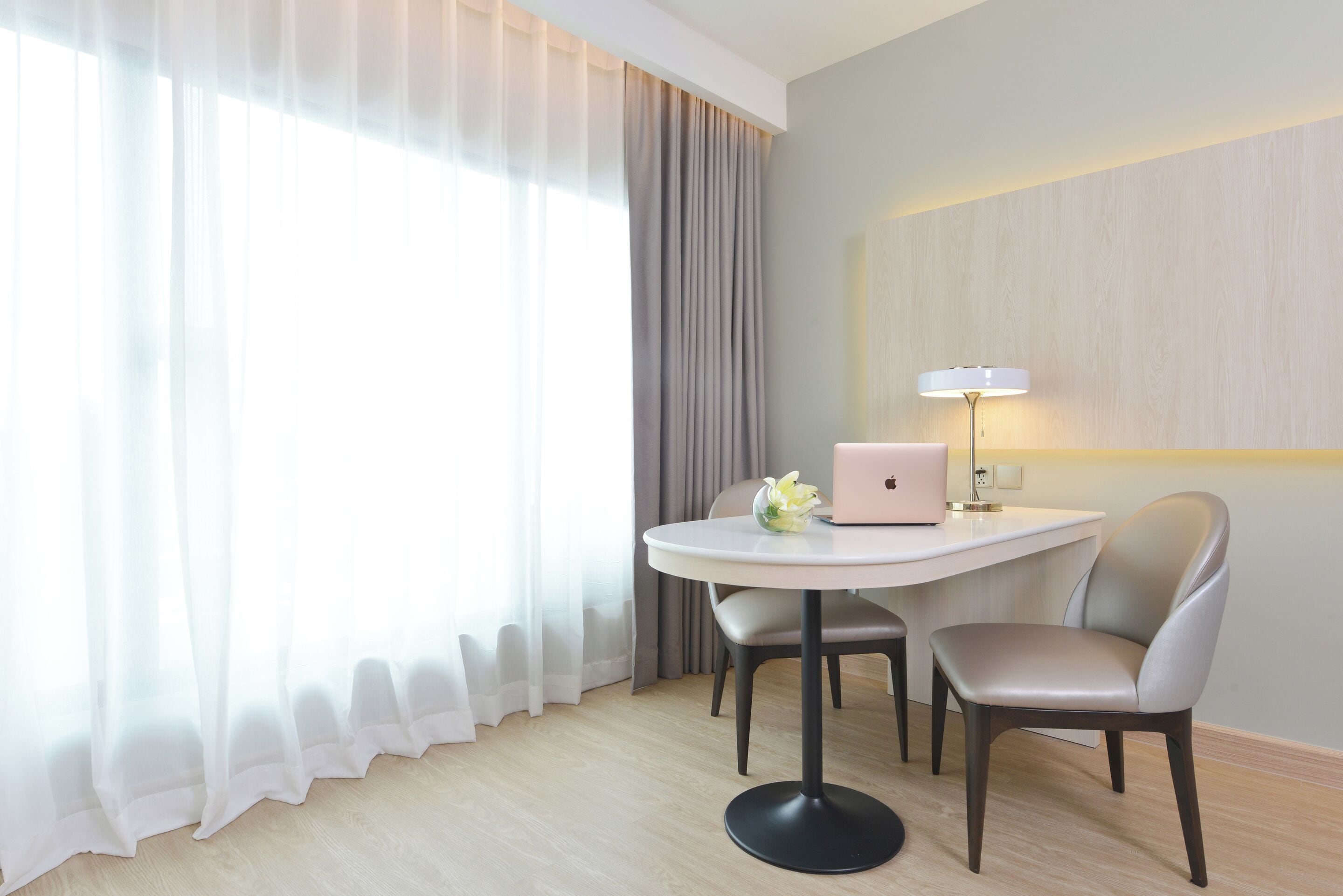 A hotel room with a round table, two chairs, a laptop, a lamp, and a window with sheer curtains.