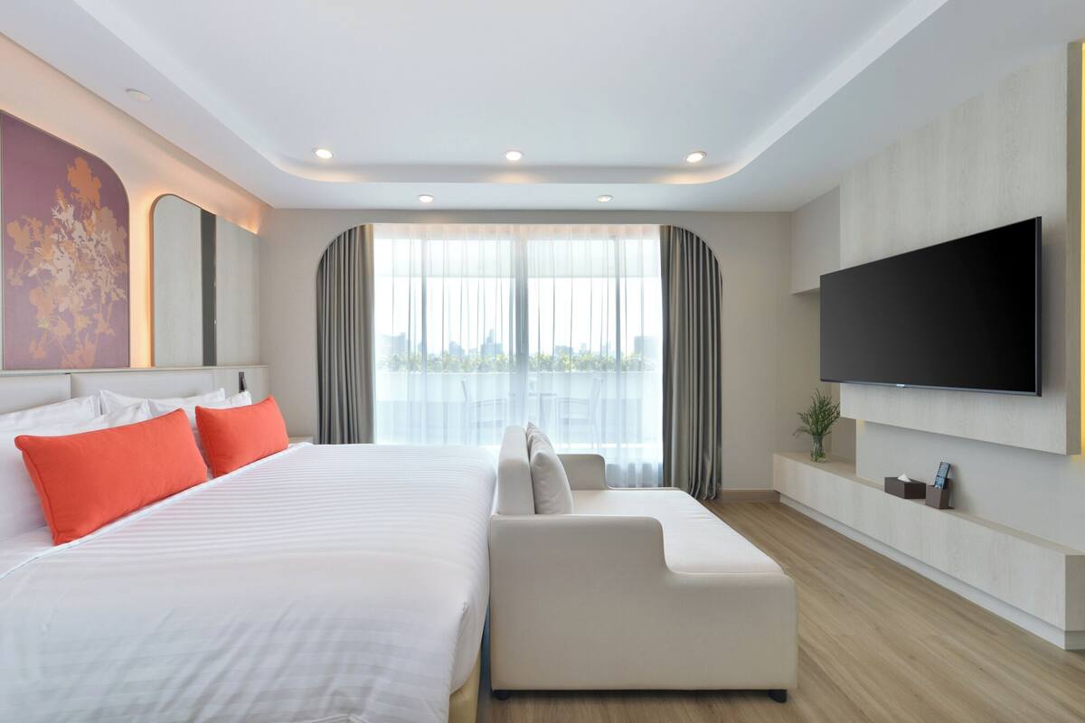 A modern hotel room with a large bed, a sofa, a flat-screen TV, and a city view.