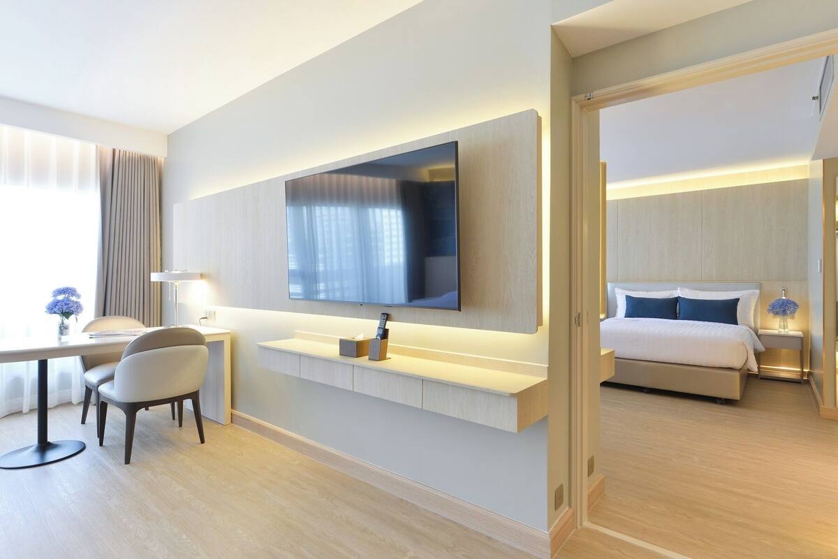 A modern hotel room with a large flat-screen TV mounted on the wall, a bedside table with a lamp, a bed with white bedding, and a desk with a chair.