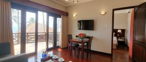 A hotel room with a dining area, a TV mounted on the wall, and a balcony with a view of the beach.