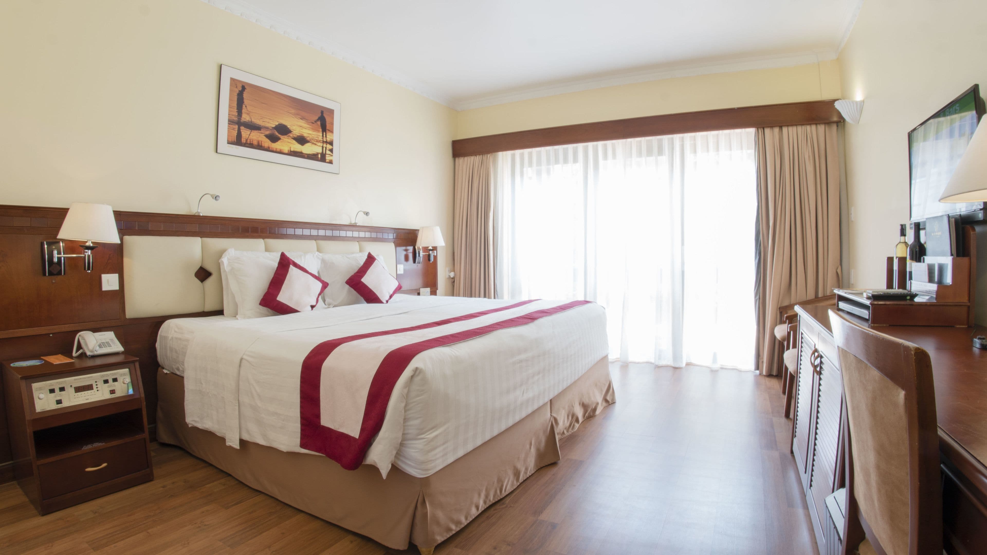 Una camera d'albergo con un letto grande, una testiera in legno, un comodino con una lampada, un tavolino con il telefono e una scrivania con un computer, oltre a una finestra con tende.