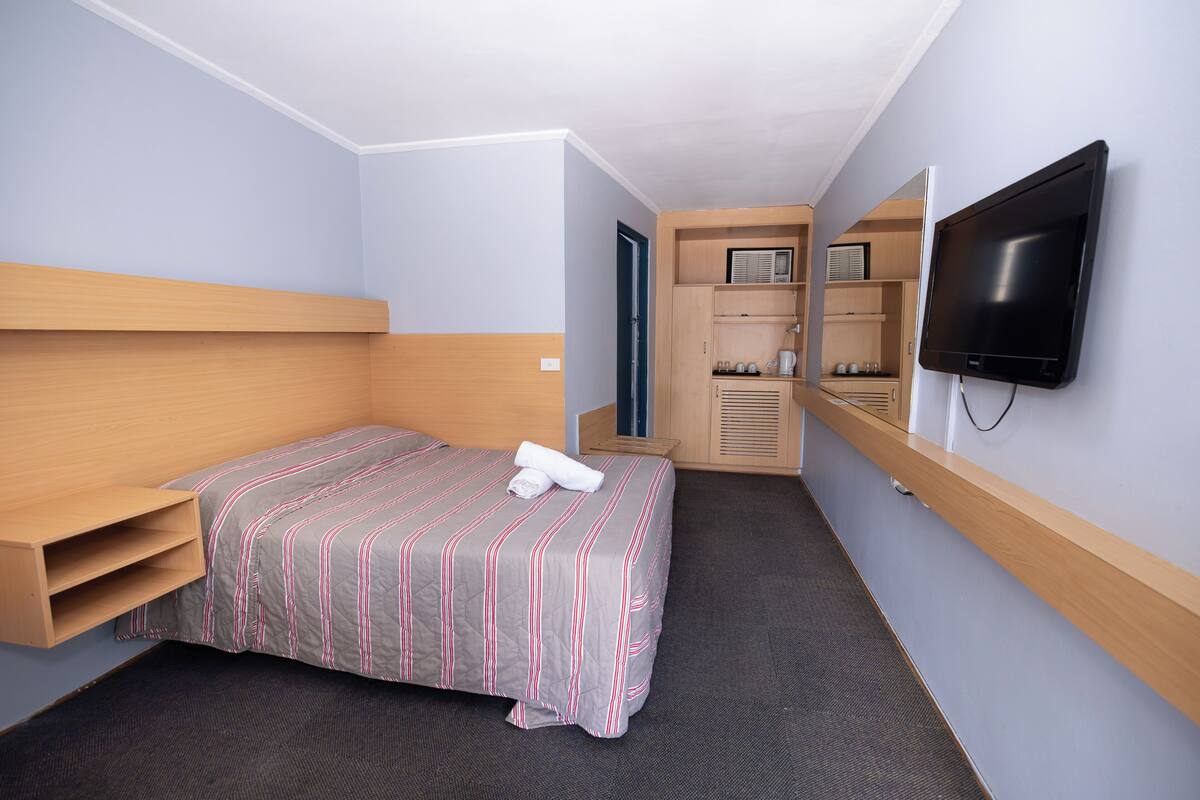 Una habitación de hotel con cama, televisión, estante de madera y una pequeña kitchenette.