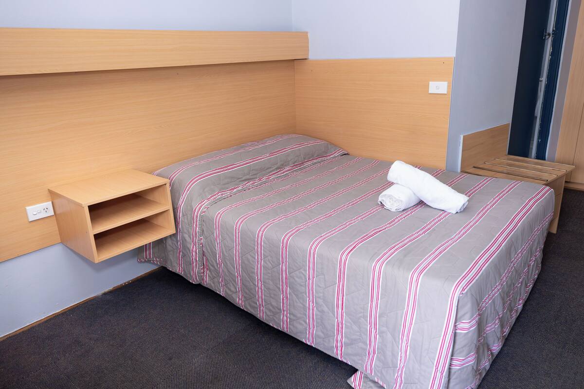 Una cama individual con una colcha a rayas, un cabecero de madera, un pequeño estante y una puerta a la derecha.