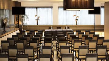 Un salón de conferencias con filas de sillas, una mesa larga y ventanas amplias con cortinas.