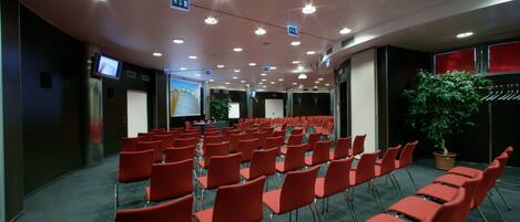 A conference room with rows of red chairs, a large screen, and a projector.
