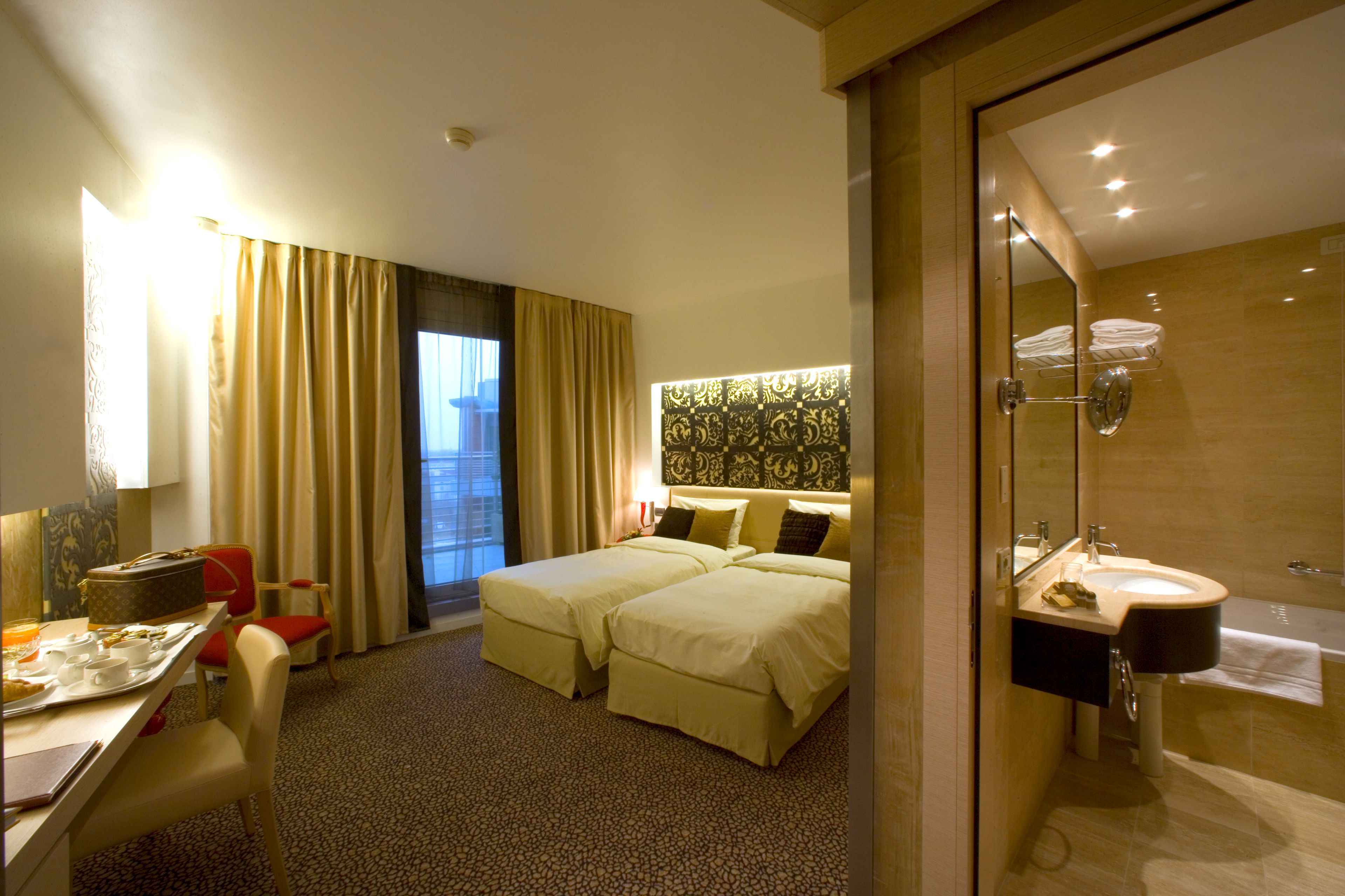 Una camera d'albergo con un letto, una scrivania con una sedia, un bagno con lavandino e specchio, e una finestra con tende.