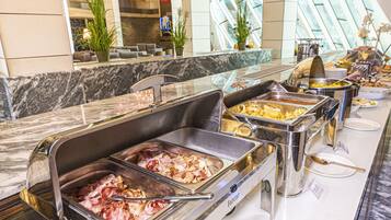 Un bufé con diversas opciones de comida, incluyendo carnes y frutas, en un moderno entorno hotelero con mostradores de mármol y plantas decorativas.