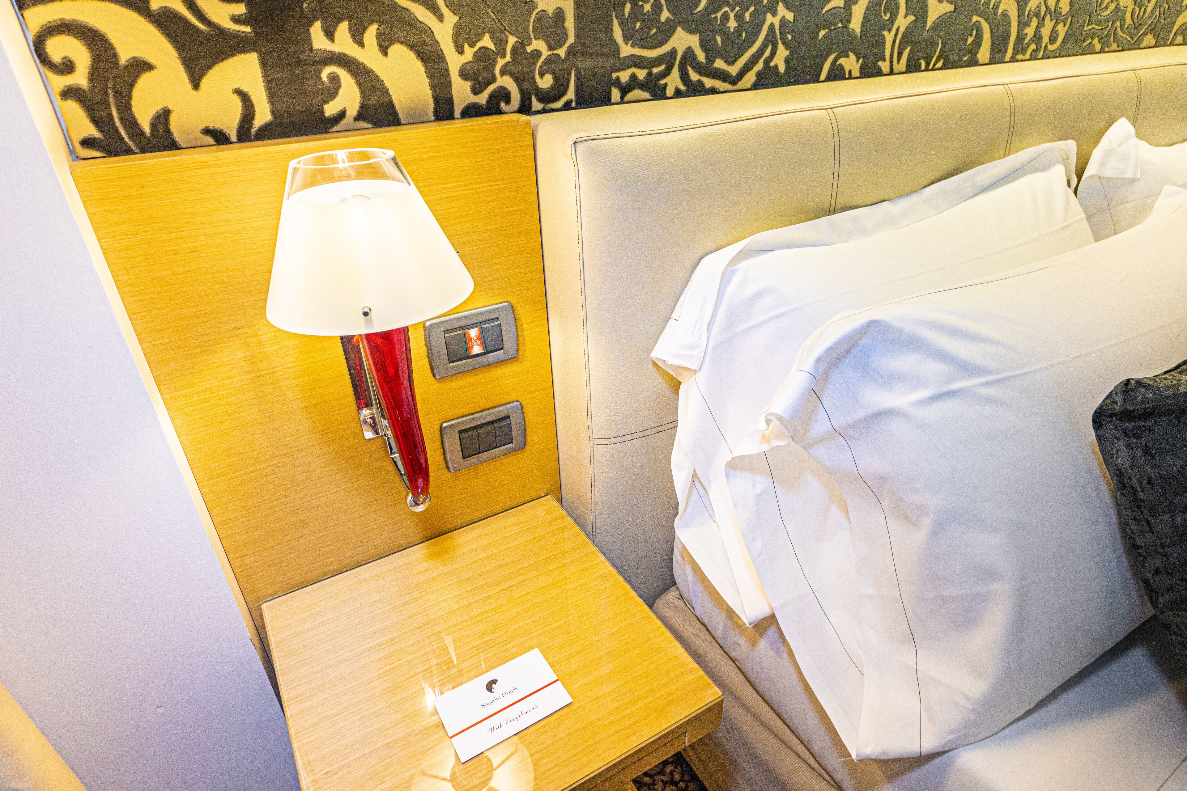 A hotel room with a wooden nightstand, a red lamp, a white bed with pillows, and a decorative wall pattern.