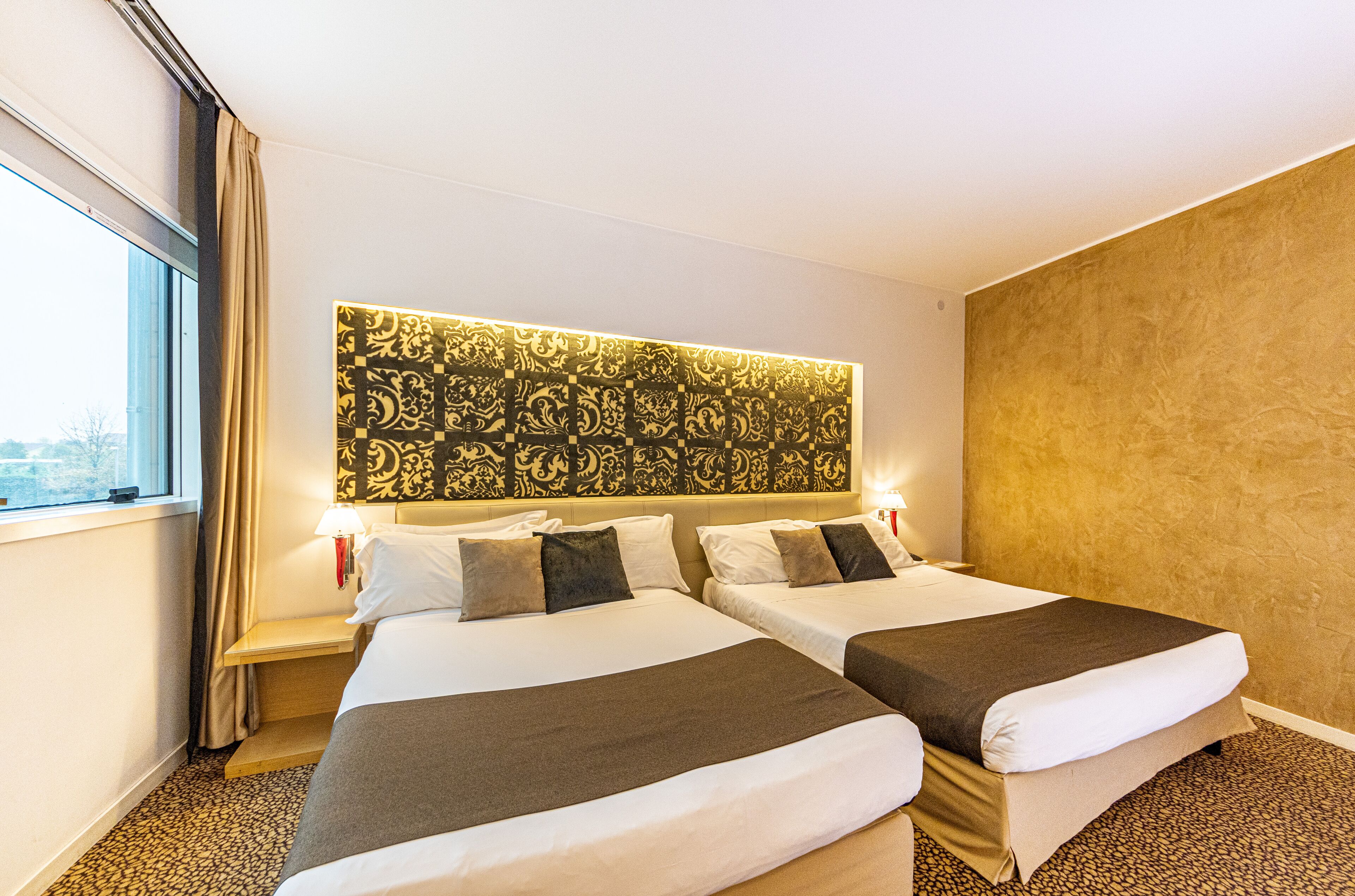 A hotel room with two beds, a patterned headboard, bedside lamps, and a view of the city through the window.