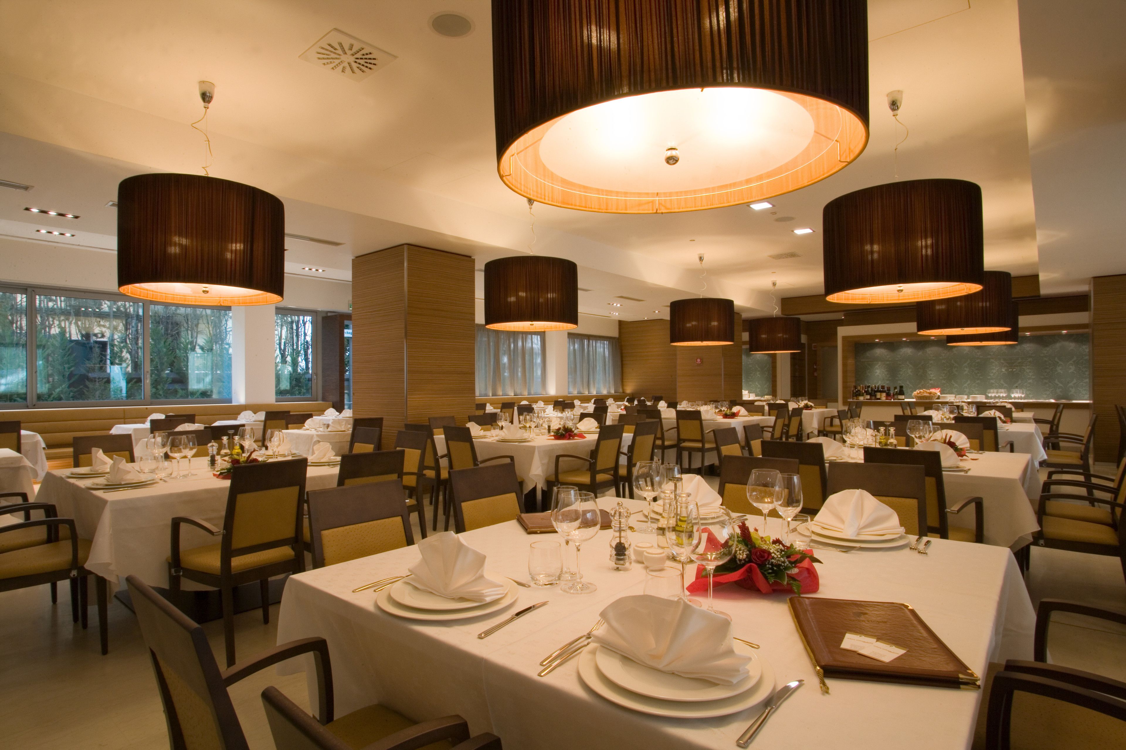 Un restaurant doté de nombreuses tables dressées pour le service, avec des nappes blanches, des couverts disposés avec soin et un éclairage d’ambiance.