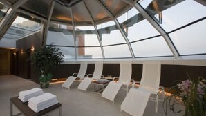 A modern lounge area with white lounge chairs and a table with towels, under a glass ceiling with a view of the sky. - Antony Palace Hotel (Marcon)