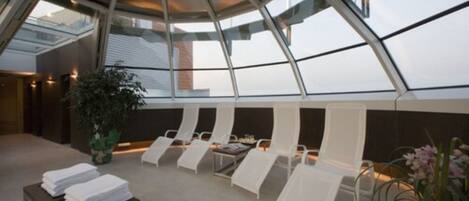 Un espace de détente moderne avec des fauteuils blancs et une table où sont disposées des serviettes, le tout sous un plafond de verre offrant une vue sur le ciel.