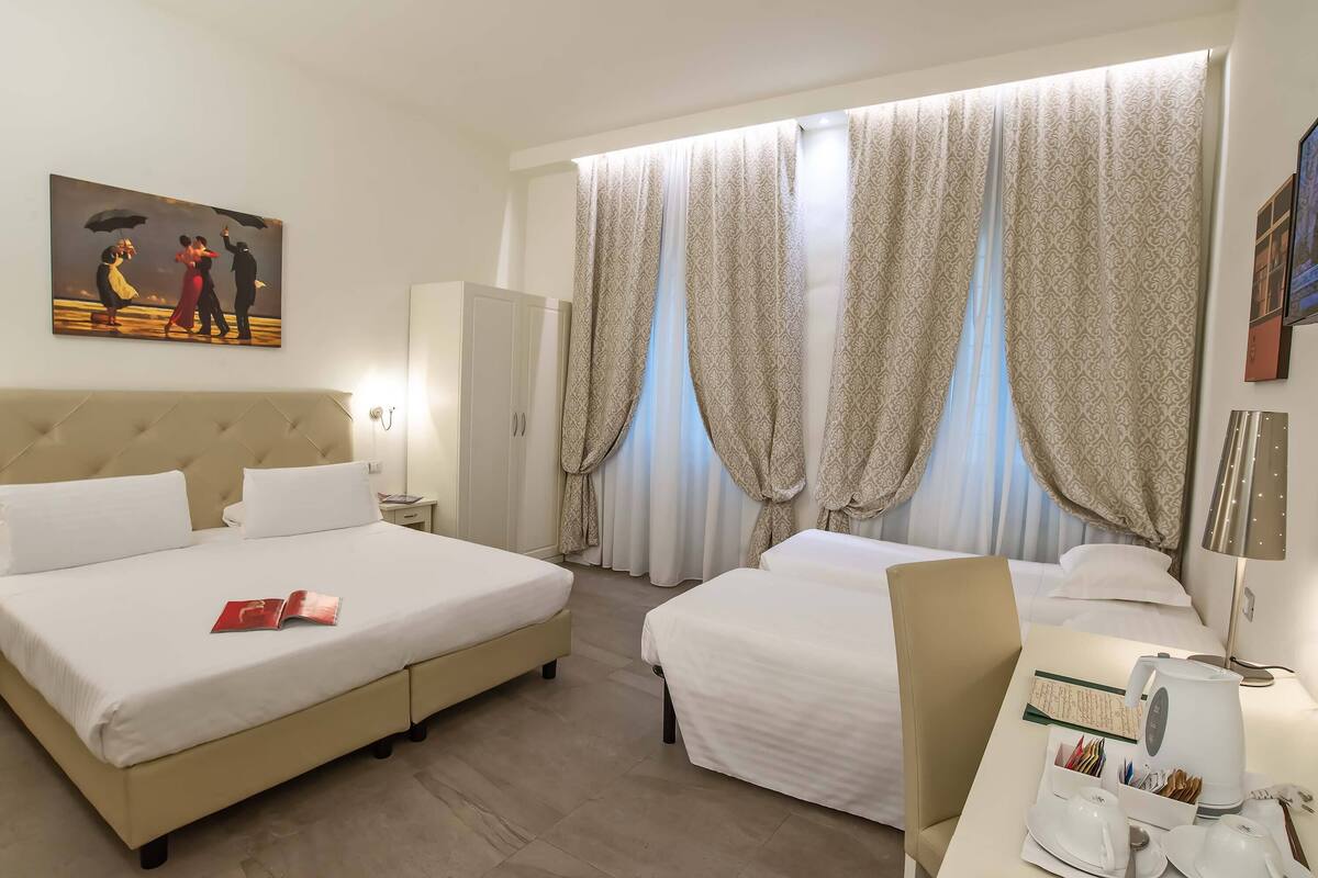 A hotel room with a large bed, two smaller beds, a desk with a kettle, cups, and a book.