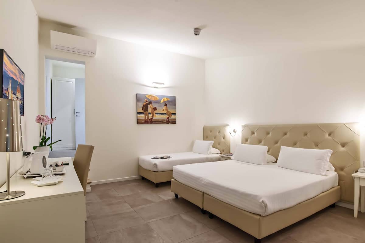 A hotel room with two beds, a desk with a lamp, a painting on the wall, and an air conditioning unit.