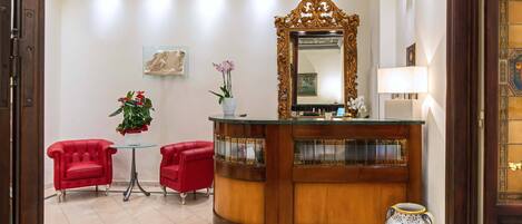 A reception area with a wooden reception desk, two red armchairs, a small table with a potted plant, a large ornate mirror, and a decorative vase.