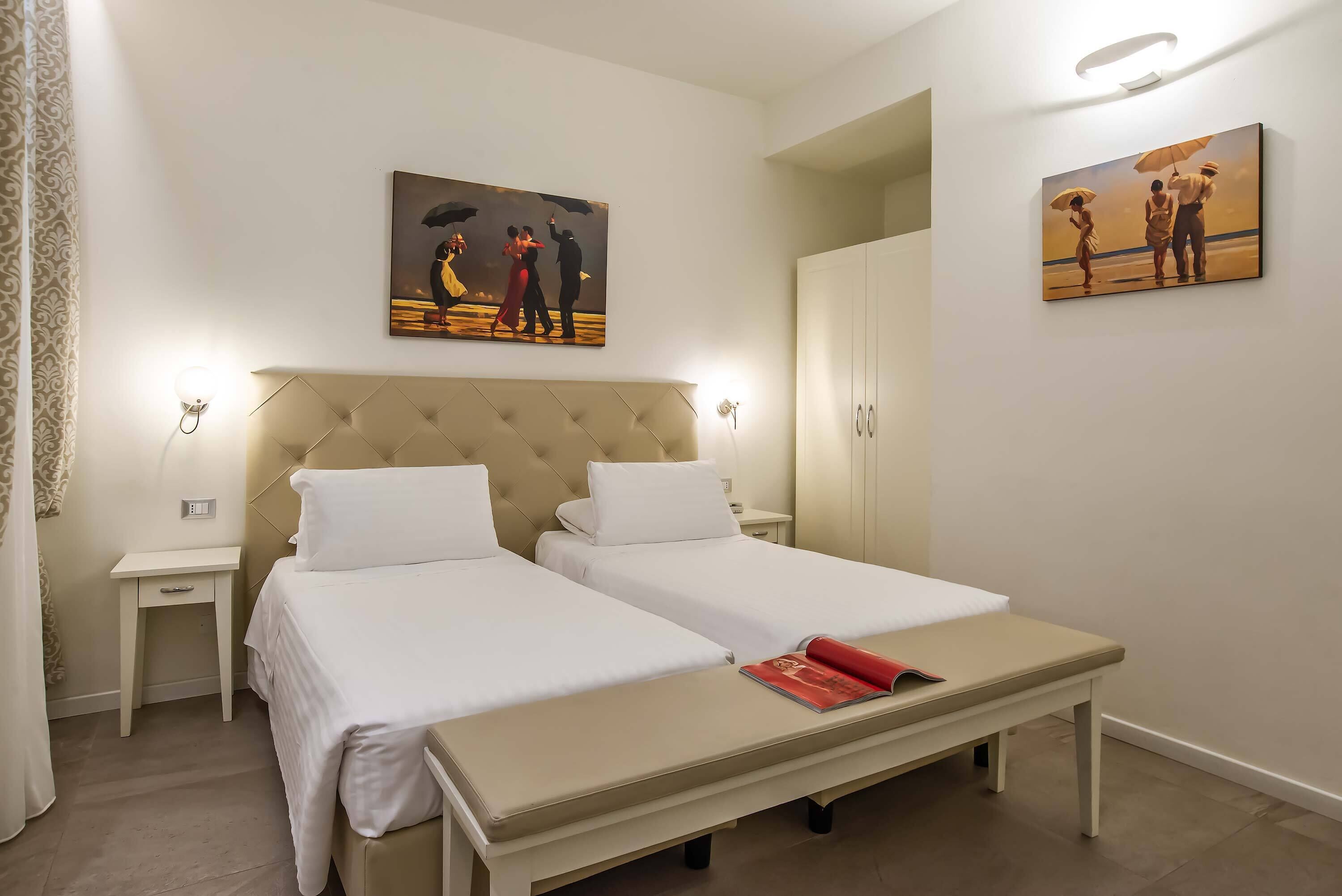 A hotel room with two beds, a bedside table, a nightstand, a bench, and artwork on the walls.