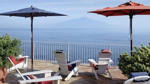 A terrace with white lounge chairs and red umbrellas, overlooking a body of water and a distant mountain. - Maison La Minervetta (Sorrento)