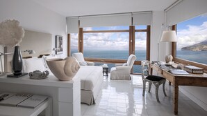 A modern bedroom with a large window overlooking the sea, a white bed, and a wooden desk with a lamp. - Maison La Minervetta (Sorrento)