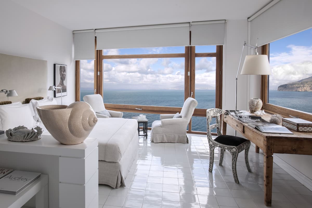 Une chambre moderne dotée d’une grande fenêtre donnant sur la mer, d’un lit blanc et d’un bureau en bois avec une lampe.