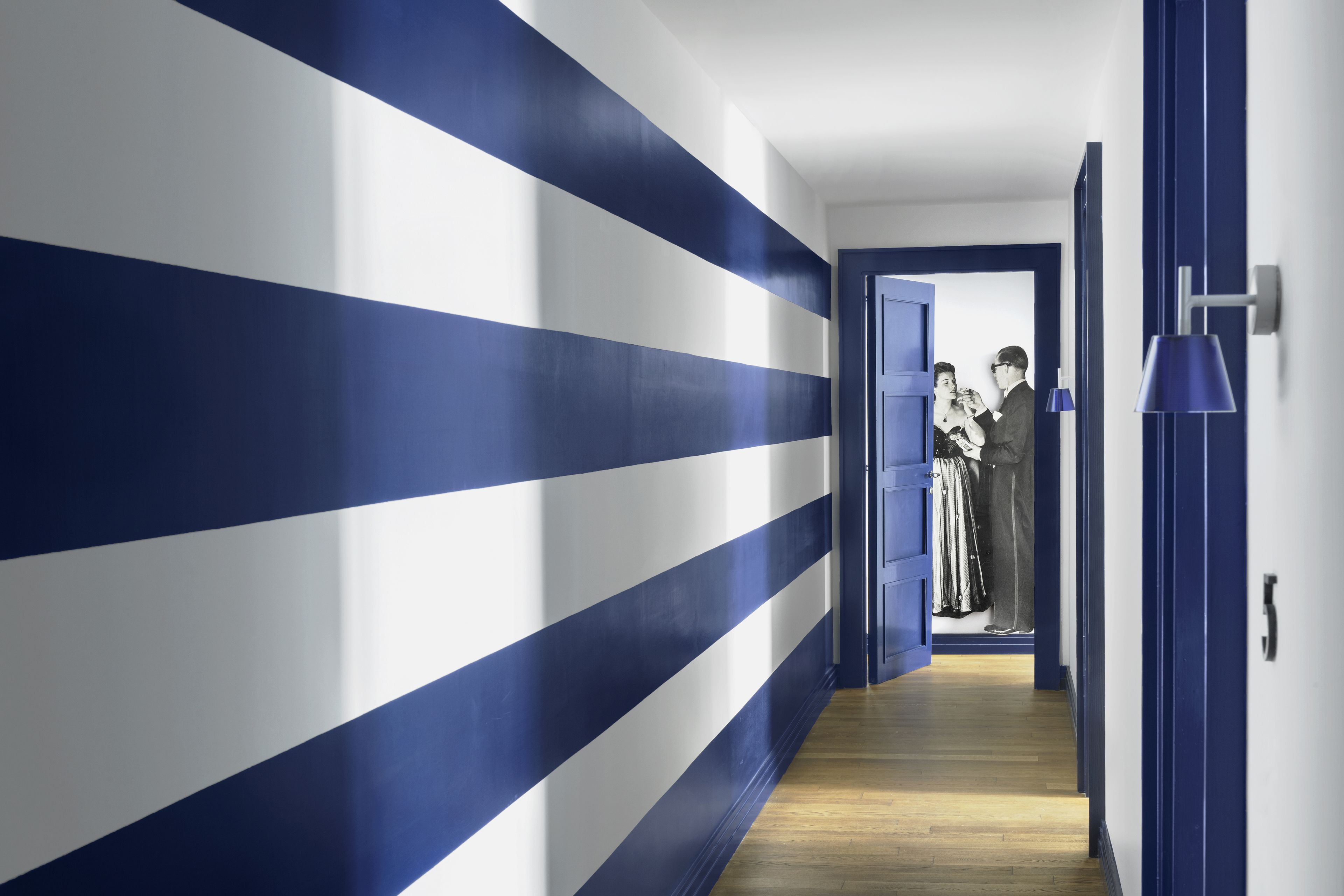 A hallway with blue and white stripes on the walls, a wooden floor, and a door leading to a room with a person in the background.