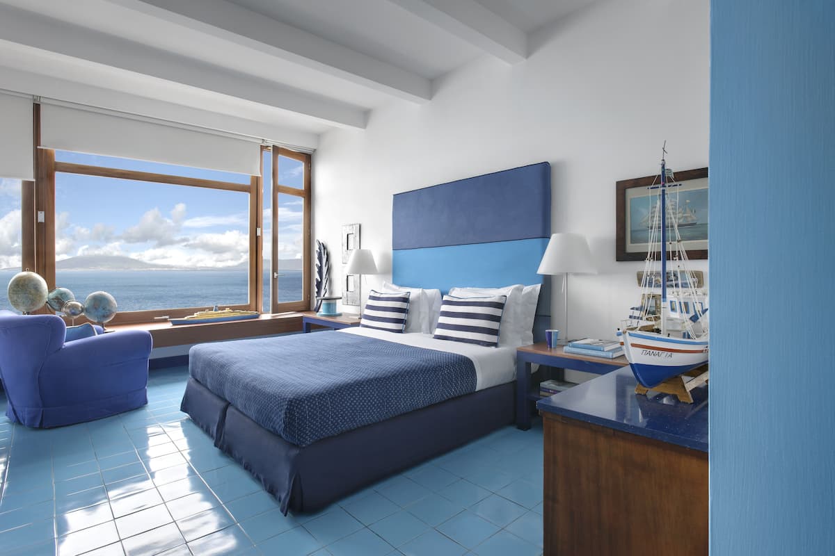 Une chambre avec un grand lit, une vue sur la mer, un fauteuil bleu et une petite table sur laquelle est posé un modèle de bateau.