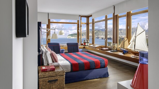 A modern bedroom with a large window overlooking a coastal view, a bed with striped bedding, two chairs, and a small table with decorative items. - Maison La Minervetta (Sorrento)