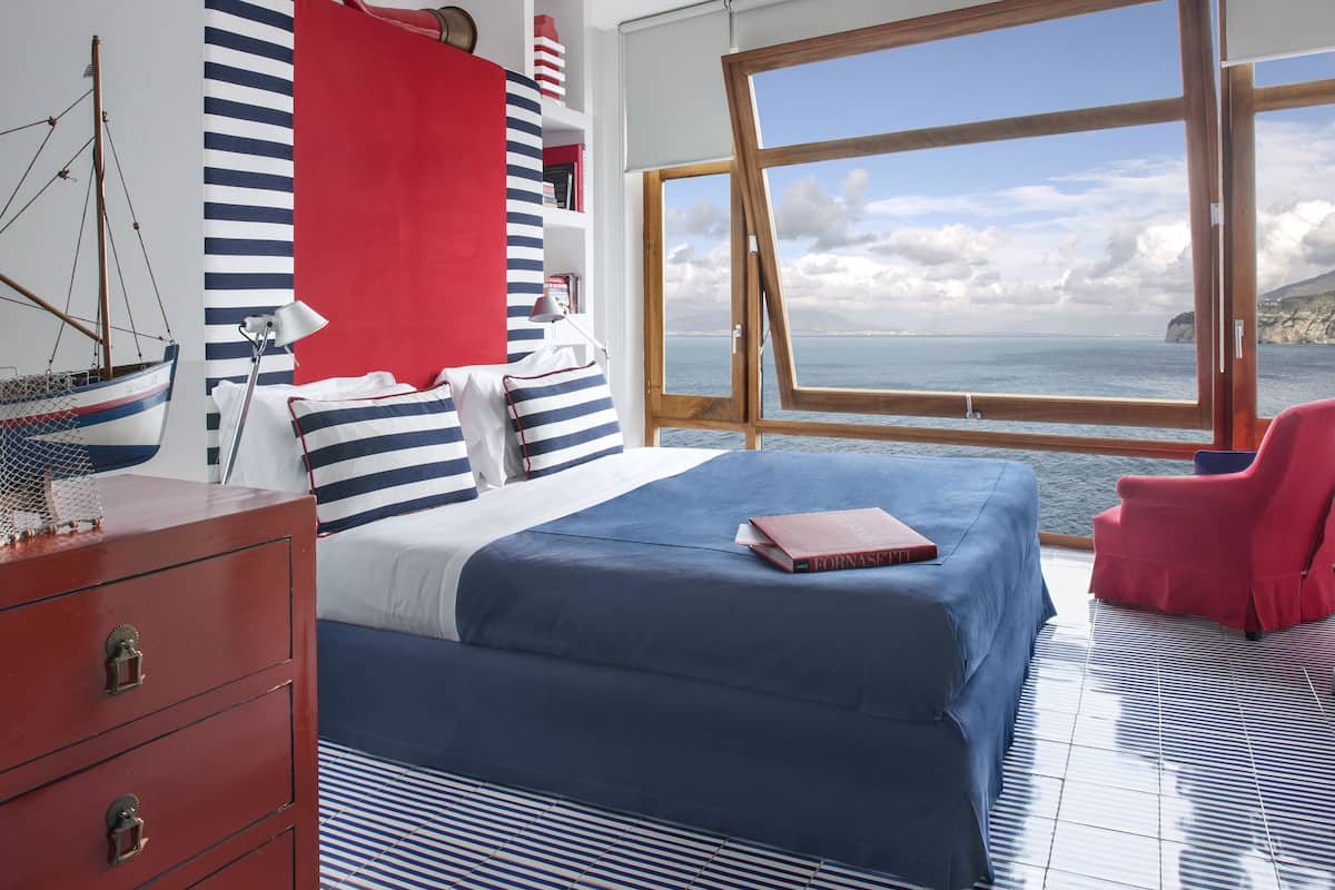 Une chambre avec une grande fenêtre, une literie à rayures, une chaise rouge et une commode en bois.