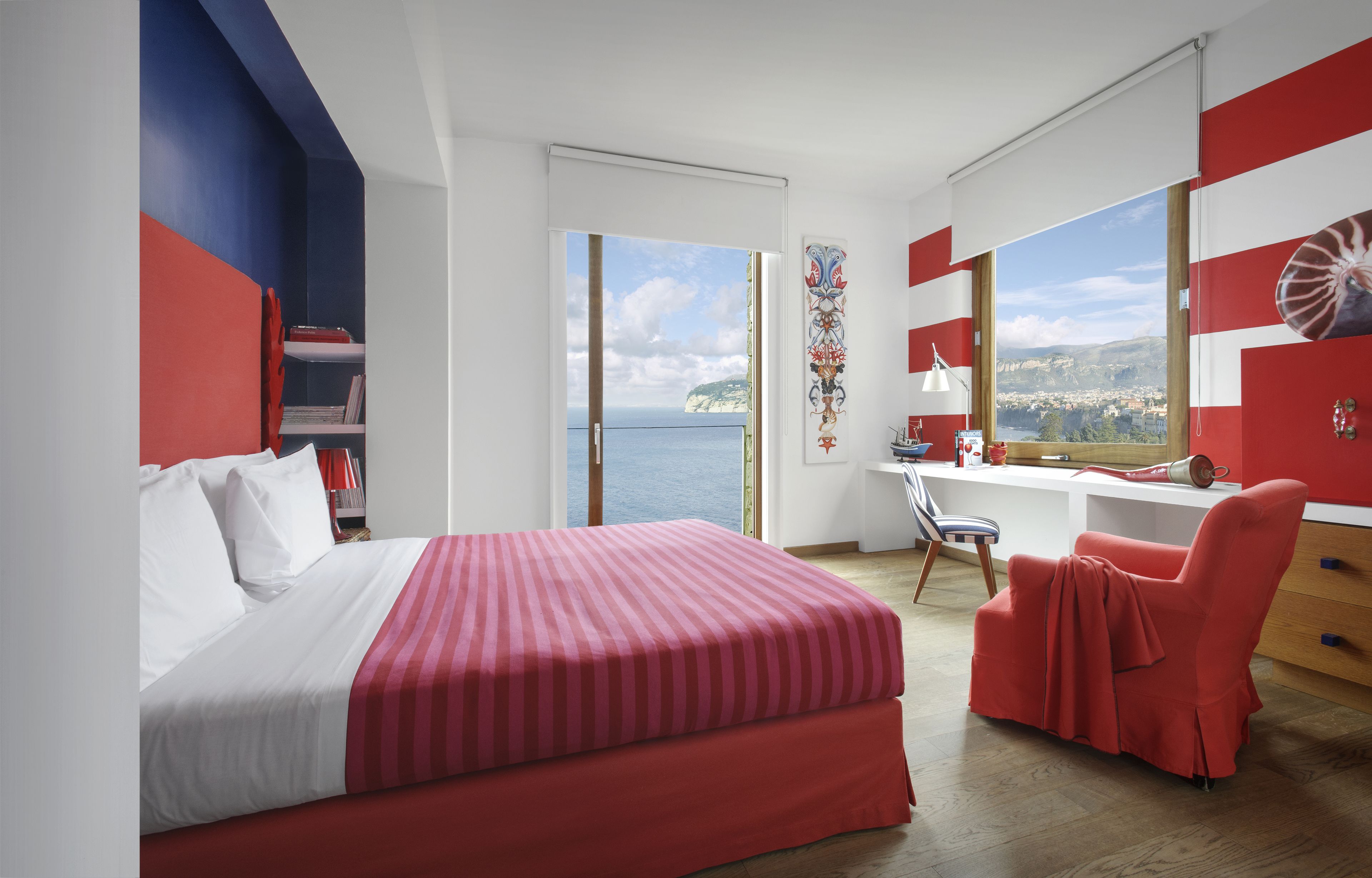 A modern hotel room with a large bed, a desk, and a chair. The room has a view of the sea and a cityscape.