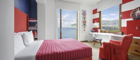 A modern hotel room with a large bed, a desk, and a chair. The room has a view of the sea and a cityscape.