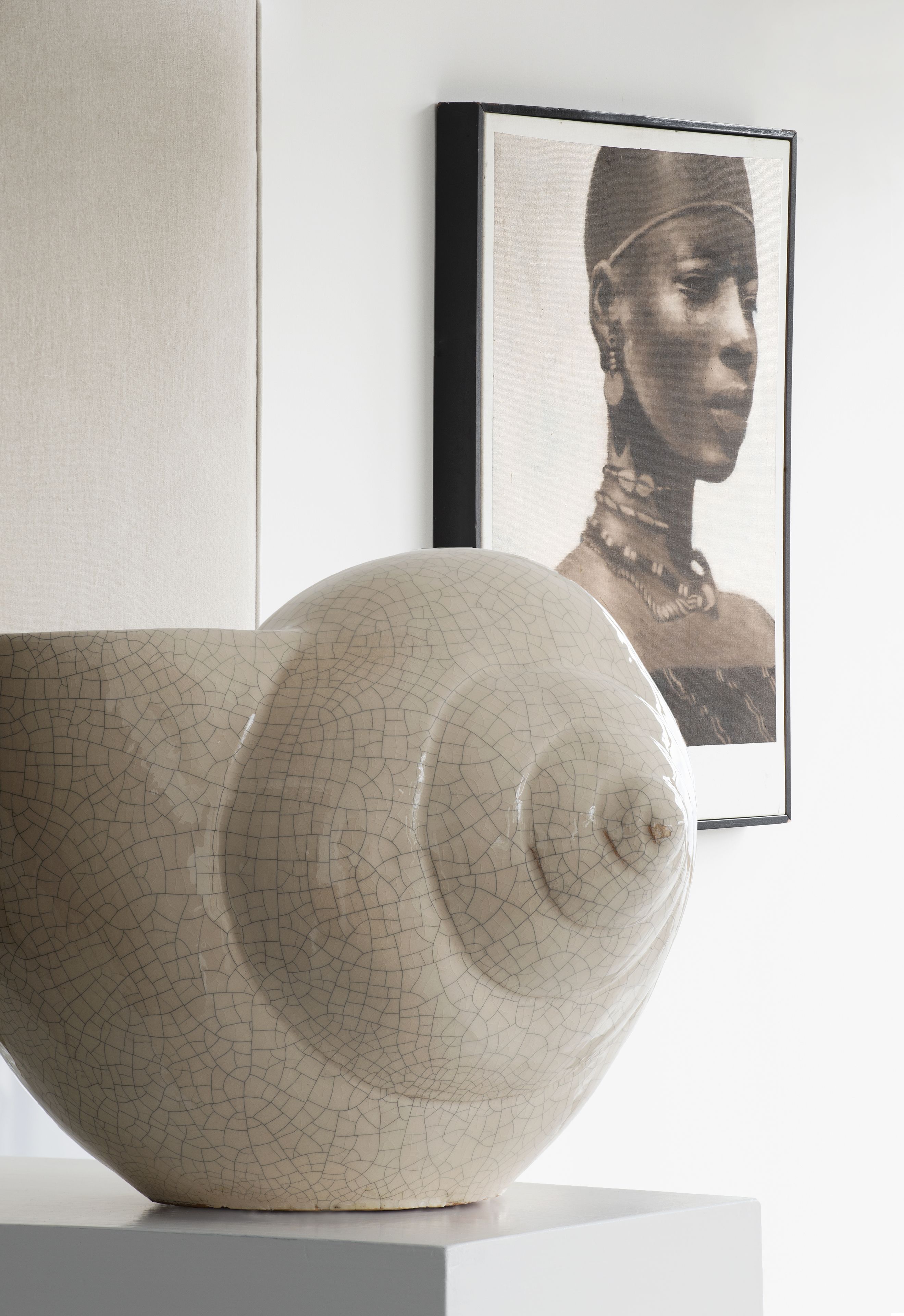 Una escultura de cerámica con textura agrietada, colocada sobre un pedestal blanco frente a una pared lisa con un cuadro enmarcado al fondo.