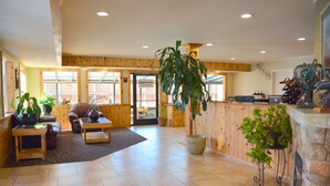 A spacious lobby with wooden flooring, a reception desk, a seating area with a sofa and armchairs, and several potted plants.