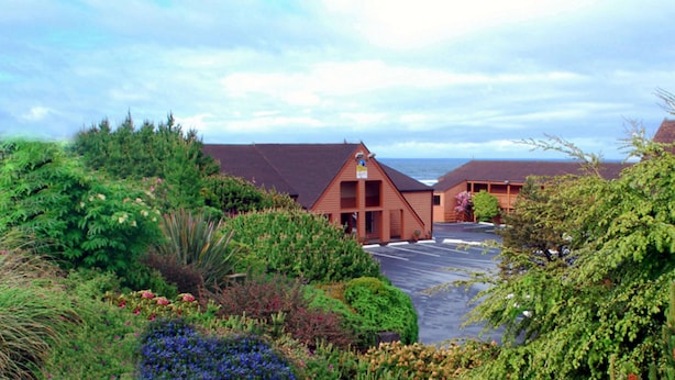 A building with a sloped roof, surrounded by lush greenery and a parking area.