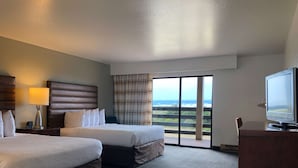 A hotel room with two beds, a TV, a lamp, and a view of the ocean through large windows.