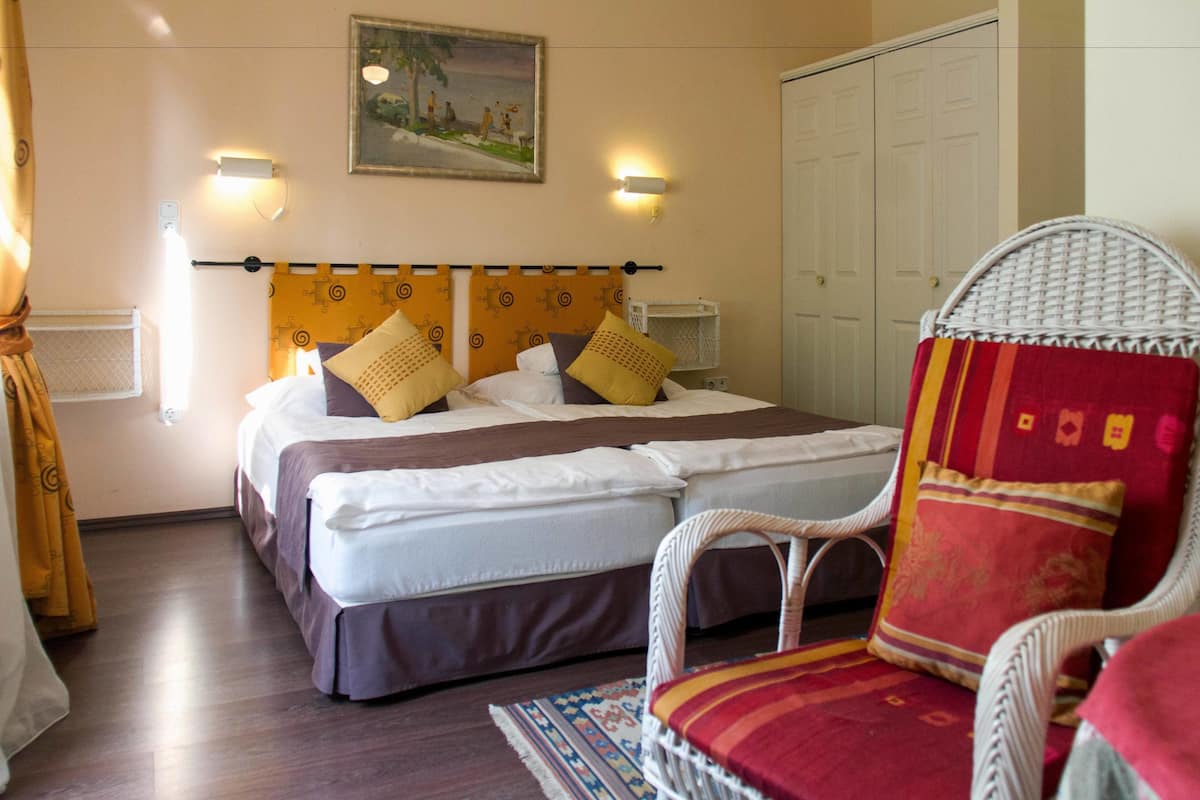 Een tweepersoonsbed met hoofdbord, twee bedlampjes, een rieten stoel met een rode kussen en een schilderij aan de muur.