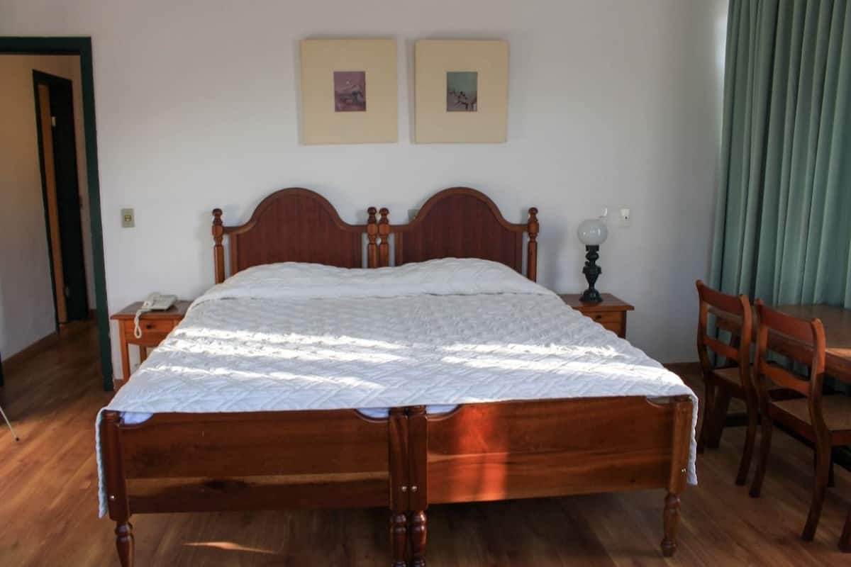 Una cama de madera con colcha blanca, una mesita de noche con lámpara y dos cuadros en la pared.