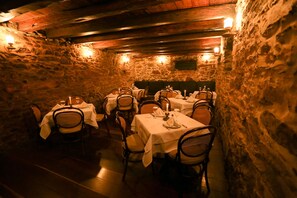 A dimly lit underground dining area with stone walls and wooden beams, featuring tables set with white tablecloths and candlelight.