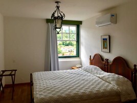 Un dormitorio con una cama de madera, una lámpara colgante, una ventana con cortinas, un aire acondicionado y un cuadro en la pared.
