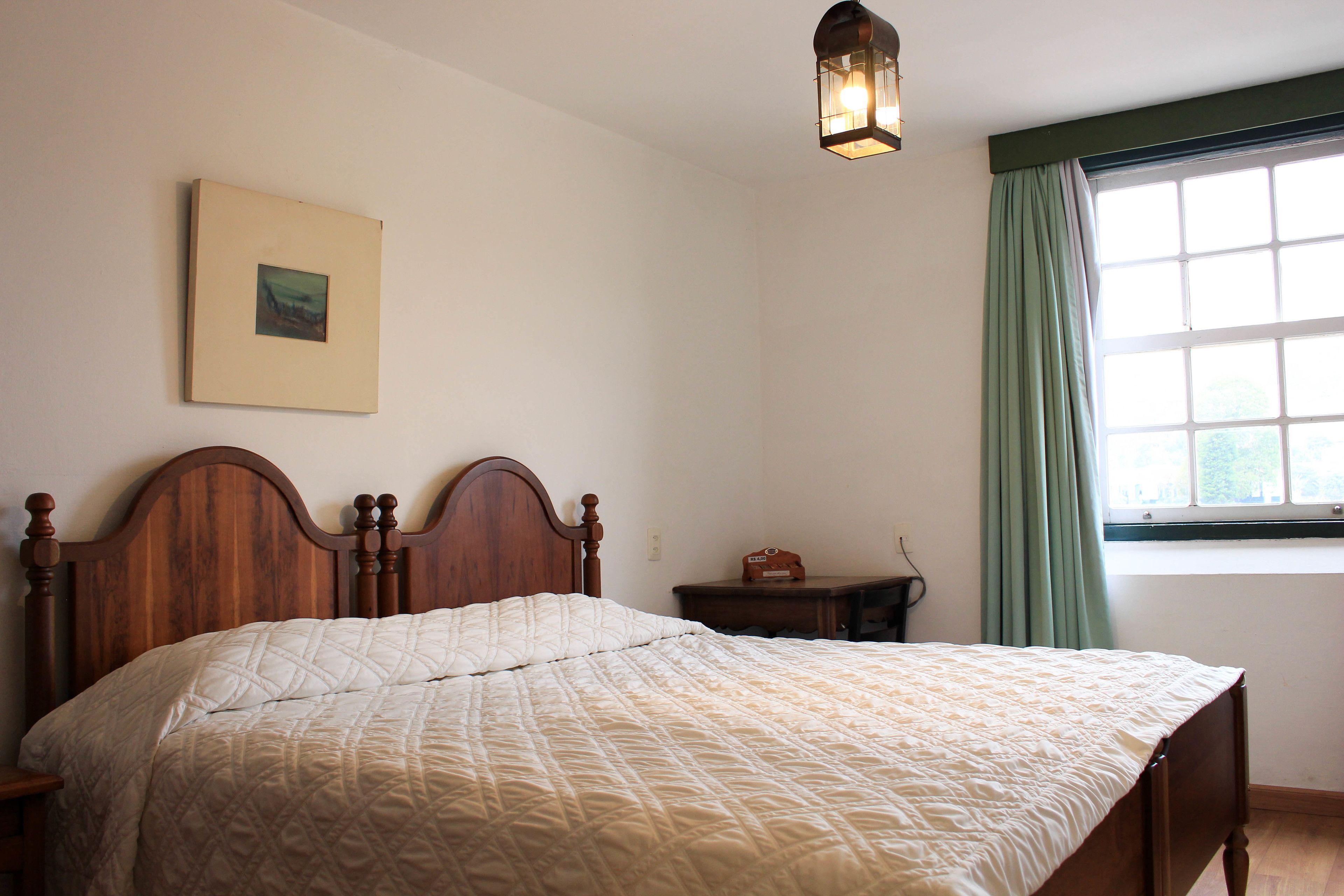 A neatly made bed with a quilted blanket, a wooden headboard, a bedside table with a lamp, and a window with curtains.