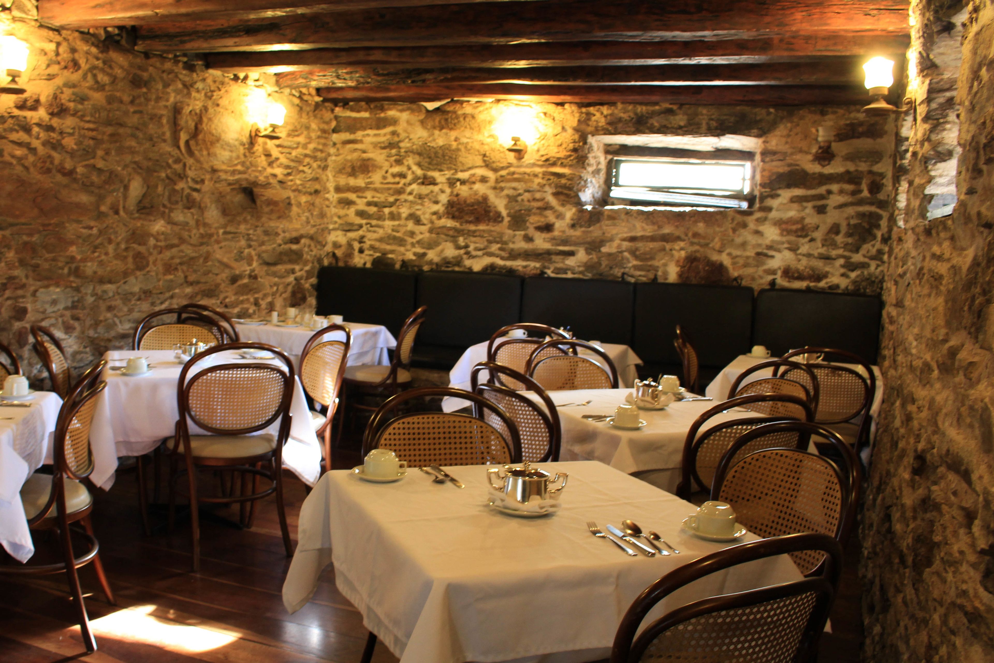 A rustic dining area with stone walls, wooden beams, and white tablecloths. Tables are set with cutlery and teapots.