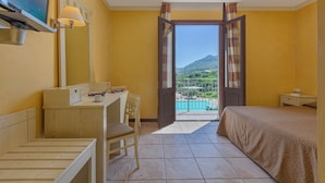 A hotel room with a desk, chair, bed, and a large window with a view of a mountain and pool.
