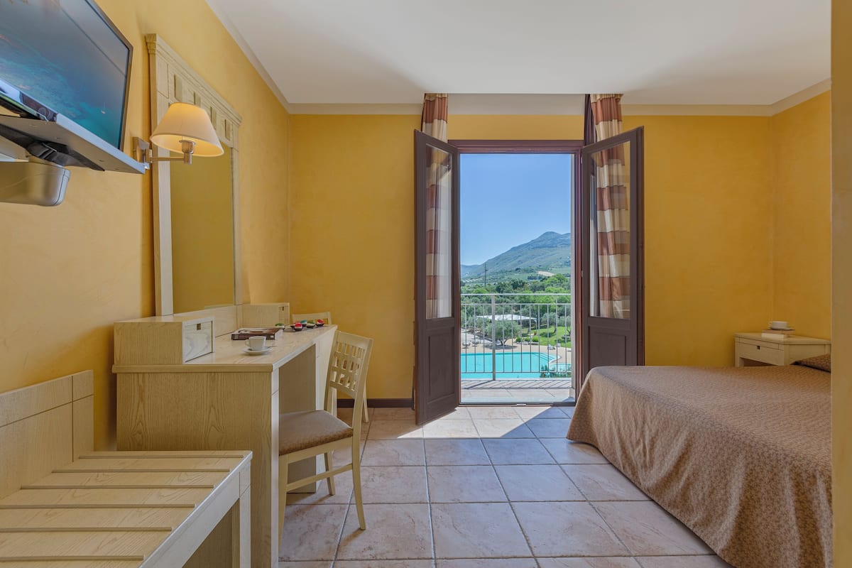 Camera d'hotel con scrivania, sedia, letto e un'ampia finestra che offre vista su una montagna e una piscina.