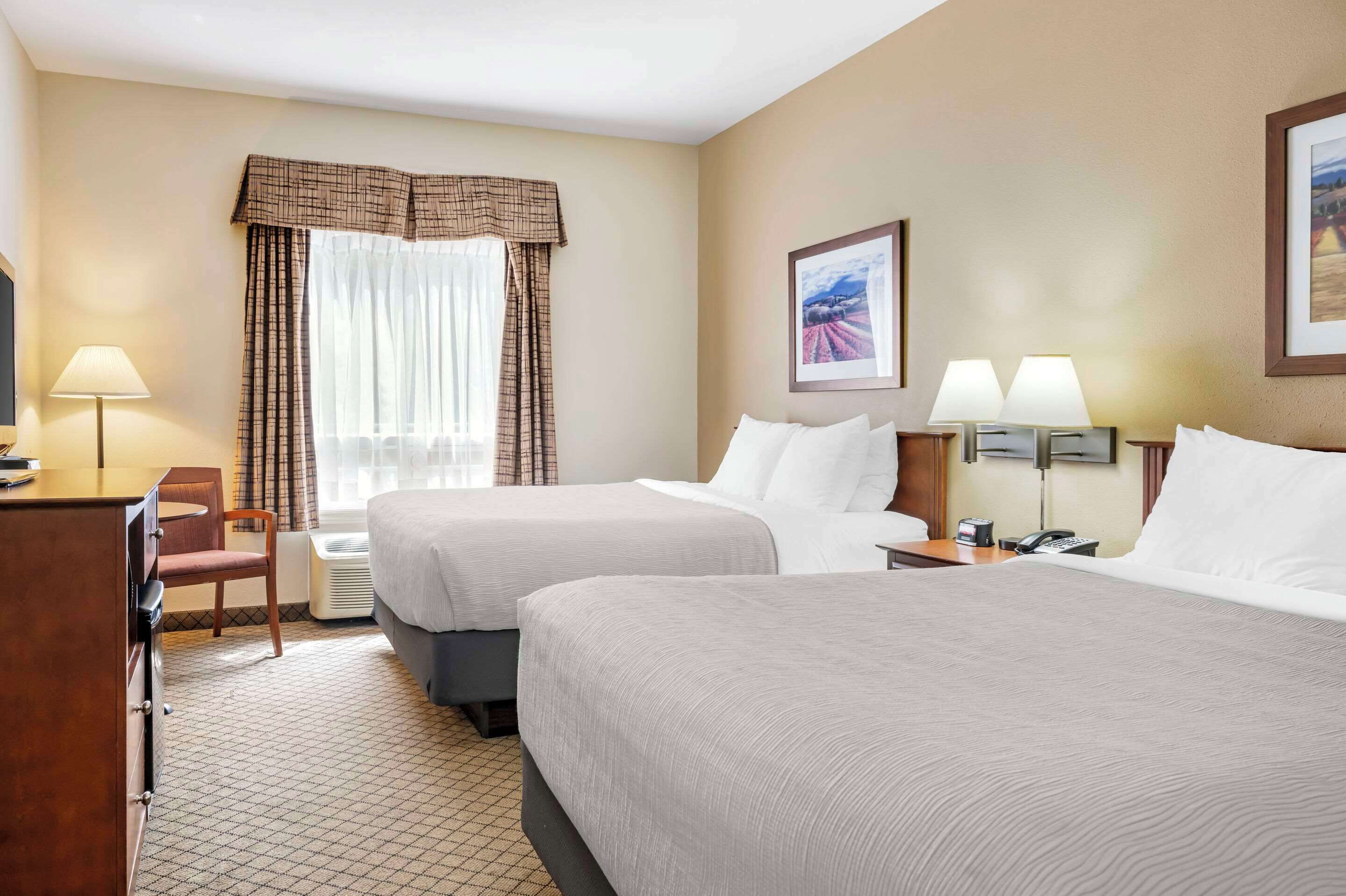 A hotel room with two beds, a desk, a chair, a lamp, and a framed picture on the wall.